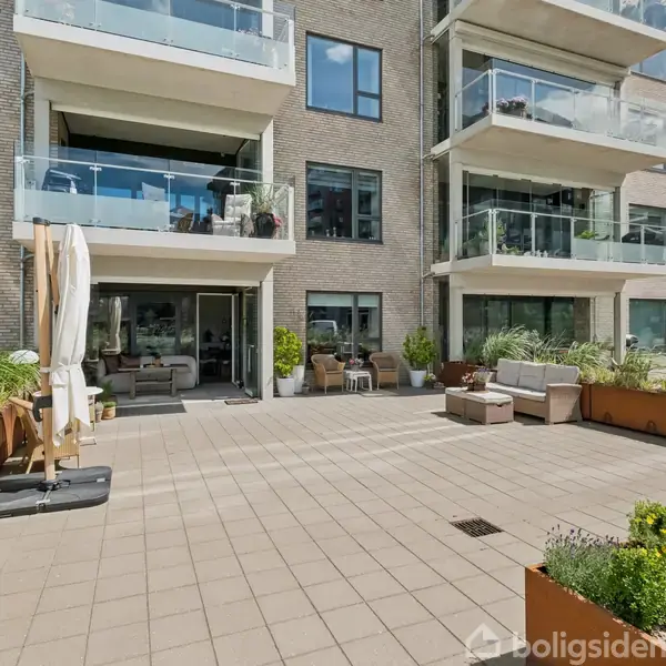 En rummelig terrasse med fliser indeholder havemøbler og planter i krukker. Store altaner og vinduer fra en moderne boligbygning omkranser terrassen, skaber en lys og åben atmosfære.