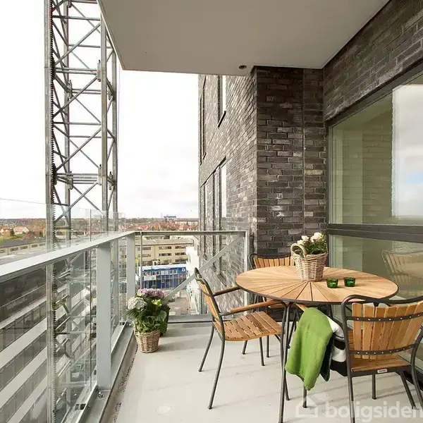 En rund trægård med fire stole står stille på en moderne lejlighedsbalkon. Omgivet af byudsigt, har bordet et par planter og lys, mens en struktur ses i baggrunden.