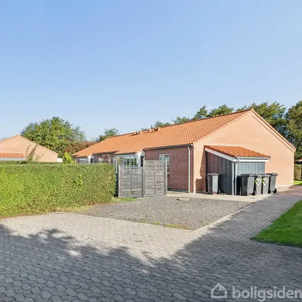 Rækkehus med rødt tegltag står i solskin ved siden af en flisebelagt indkørsel. Flere skraldespande ses ved siden af huset, omkranset af grønne buske og træer.