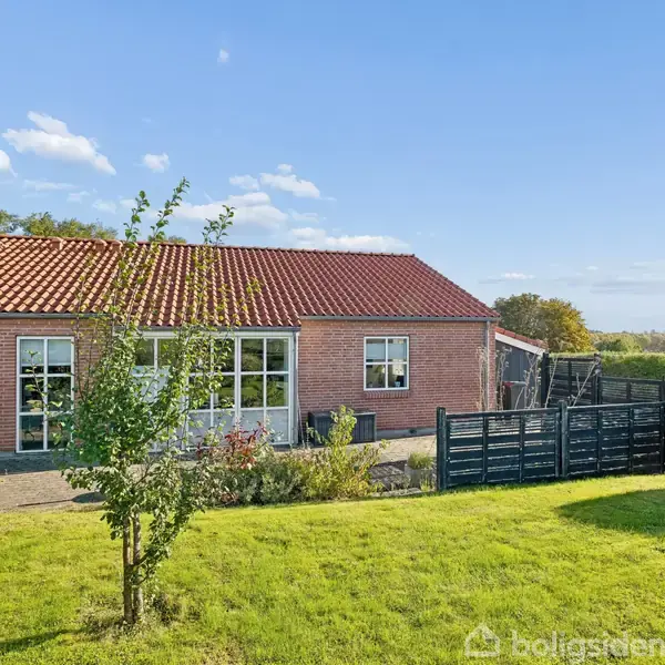 Rødstenshus med rødt tegltag har en velplejet have. Foran huset er der en grøn græsplæne med spredte buske og træer. I baggrunden er der blå himmel og buske.