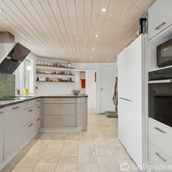 Køkken med grå skabe og skuffer, arrangere i et U. Køkkenet indeholder en emhætte over en kogeplade, en mosaik flise væg, hylder med krukker samt indbyggede apparater.