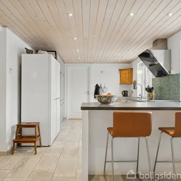 Køkken med to barstole ved høj bar, moderne udstyr og lyse farver. Træloft med indbyggede spotlights og stort vindue til venstre. Skabe og apparater i baggrunden.