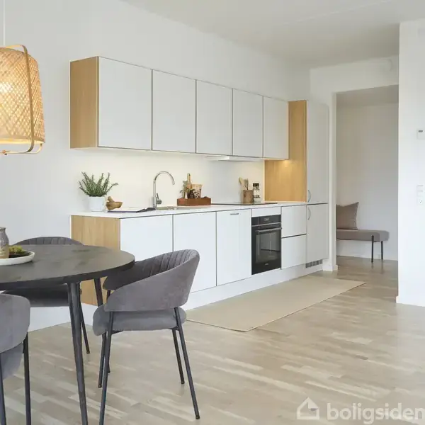 Et moderne køkken med hvide skabe og trædetaljer. En rund bordplade med stole står under en hængende lampe. Rummet er lyst med minimalistisk indretning.
