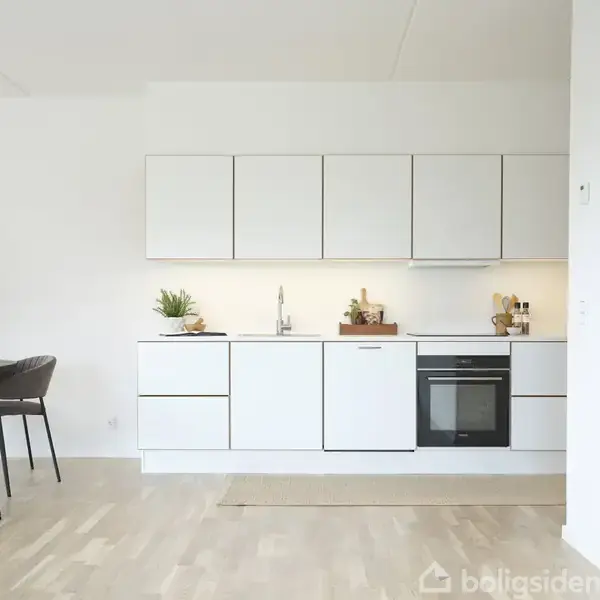 Køkken med hvide skabslåger og indbygget ovn; ved siden af står et rundt spisebord med fire stole. En dekorativ lampe hænger over bordet, og en grafisk kunstplakat ses på væggen.