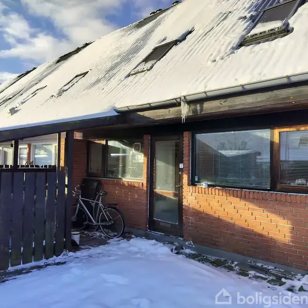 Et rækkehus med sne på taget og is på tagrenden står i et vinterligt miljø. En cykel står op ad husets røde murstensvæg ved siden af vinduerne og en dør.