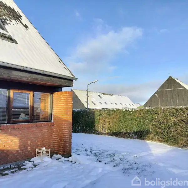 Et rødstenshus med sne på taget står i en snedækket have. Hække omkranser græsplænen, og der er klart blåt himmel.