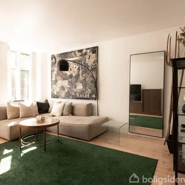 Stue med en lys sofa placeret mod en hvid væg med kunst. Rundt sofabord på grønne tæpper. Høj spejl ved siden af reol med glas og dekorative genstande.