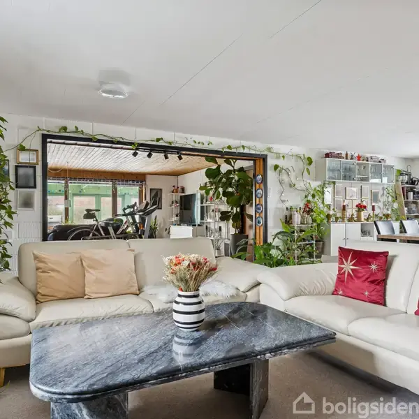 Hvid sofa med farvede puder står i en plantefyldt stue omkring et mørkt marmorbord. Store vinduer og spisebord ses i baggrunden. Ingen tekst til stede.