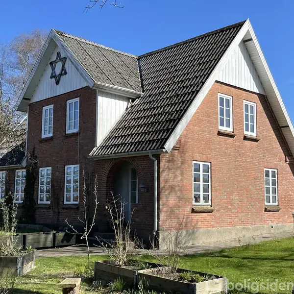 Rødt murstenshus med gavltag og stjerneornament hænger på gavlen. Huset står i en have med græs og planter omkring. Solrigt vejr.