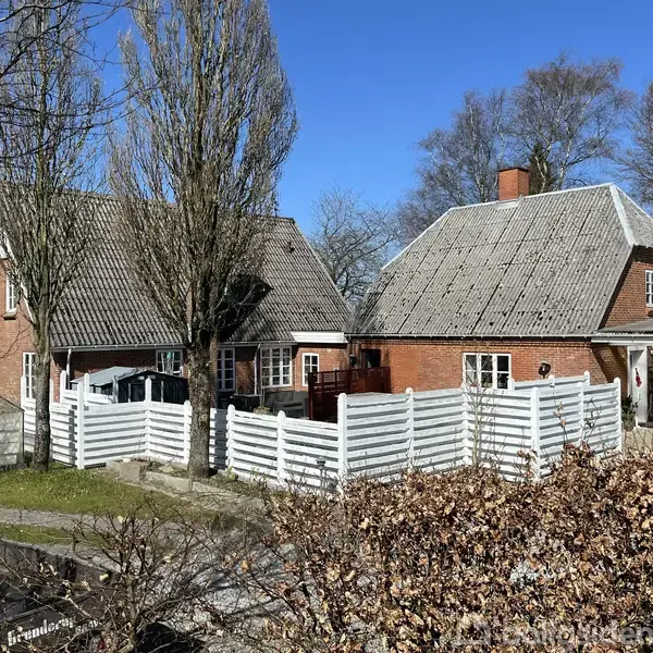 To røde murstenshuse med gråt tag er omgivet af en hvid træhegn. Et af husene har en bil parkeret under et halvtag. Omkring husene ses træer uden blade.