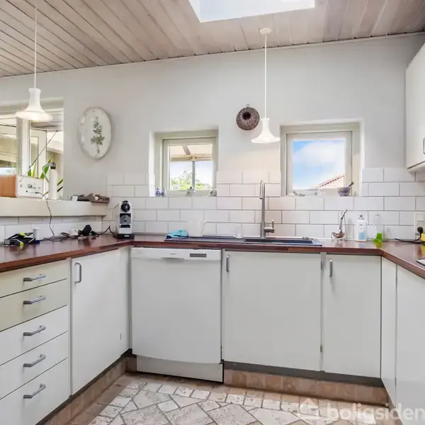Køkken med hvide skabe og træbordplader, to vinduer over vasken, flisebelagt væg, og hængelamper. Fliser på gulvet og lys træloft med ovenlys. Ingen tekst ud over "Boligsiden".