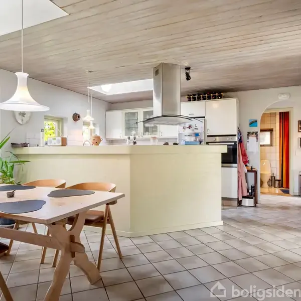 Der er et moderne køkken med hvidt spisebord og stole. Køkkenet har åbne hylder, komfur med emhætte, og store vinduer, der lader lys strømme ind. Plante i hjørnet skaber en grøn detalje.