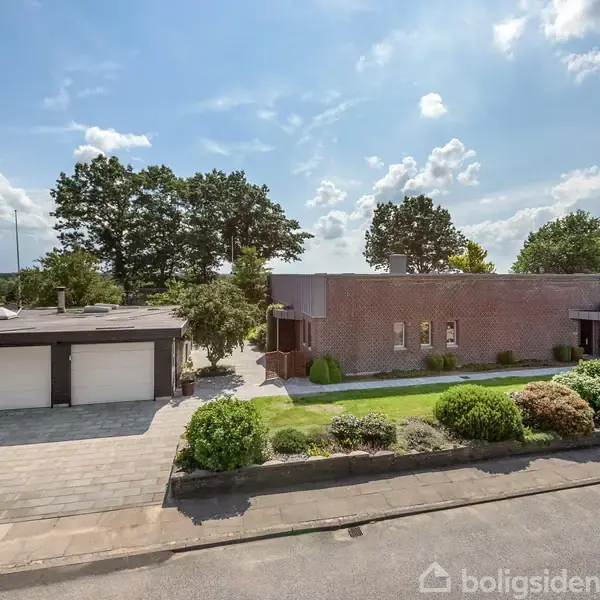 Et moderne enfamiliehus i mursten står stille i en solrig have. Ved siden af huset er en garage. Omkring huset er der buske, en velplejet græsplæne og høje træer.