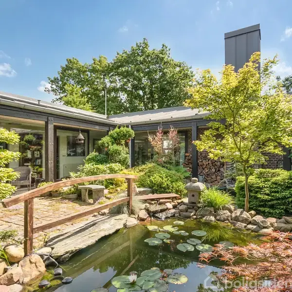 Have med blomster og dam med åkander, omkranset af stier og træer. En bro forbinder områderne, og huset ses i baggrunden under en klar blå himmel.