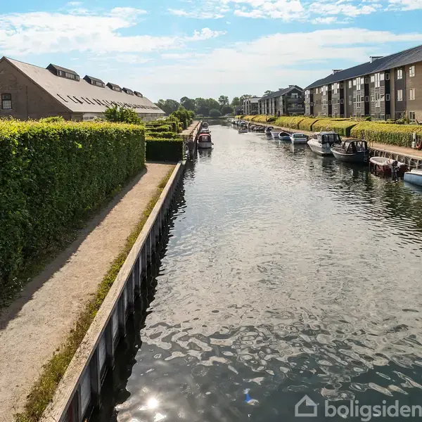 Kanal i sollys med både fortøjet langs bredden; til venstre en sti langs en hækkeindhegnet have. Omgivet af beboelsesbygninger og blå himmel.