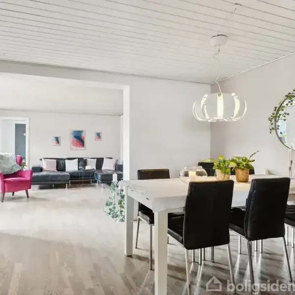 Et spisebord med sorte stole står i et lyst rum. En moderne lampe hænger over bordet. I baggrunden ses en rød stol og en sofa. En rund spejl dekorerer væggen.
