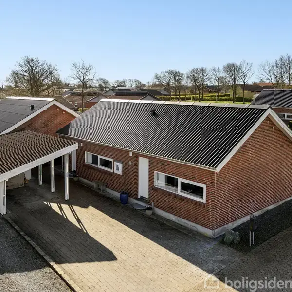 Rødt murstenshus med mørkt tag, parkeret ved carport. Beliggende i boligområde med flere lignende huse, omkranset af træer. Klar, blå himmel i baggrunden.