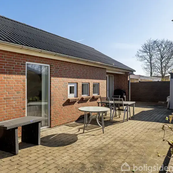 Rød murstensbygning med skydedør og små vinduer. Foran står to runde borde og en bænk på en flisebelagt terrasse i solbeskinnet have med buske og træer i baggrunden.