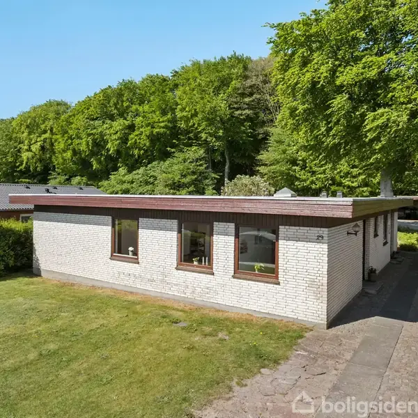 Et fladt hus med hvid murstensfacade står i en grøn have. Der er store vinduer foran, omgivet af frodig vegetation og træer i baggrunden.