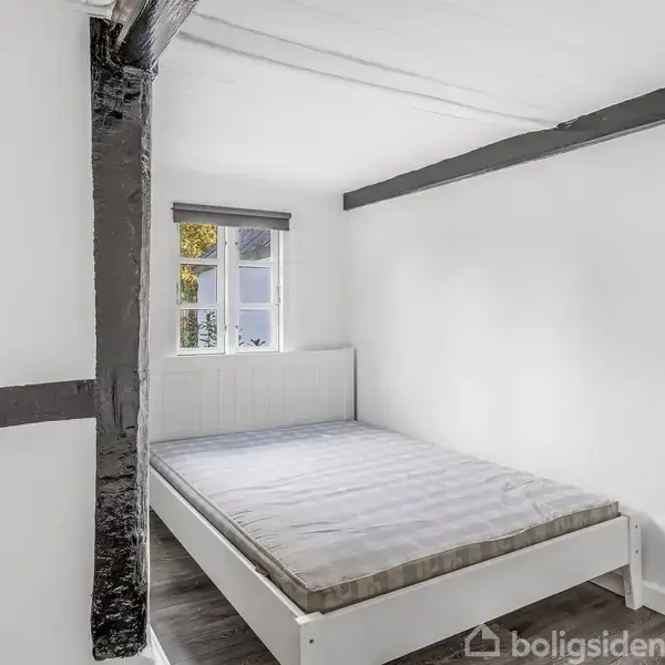 Radiator står ved hvid væg i et lille rum. Mopper, kost og tørrestativ er op ad væggen. En trætrappe fører opad tæt ved en blå dør, med lidt opbevaring langs væggen.