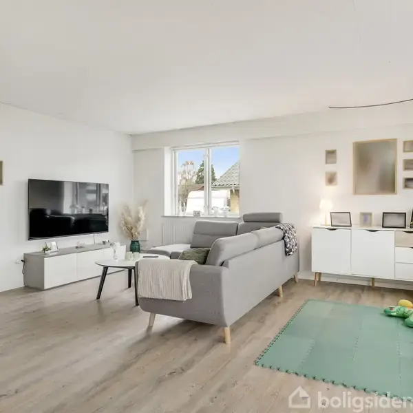 Sofa står midt i en lys stue med trælaminatgulv, flankeret af et fjernsyn på vægbordet. Ved højre væg ses et tæppe med børnelegetøj og et skab under billeder.