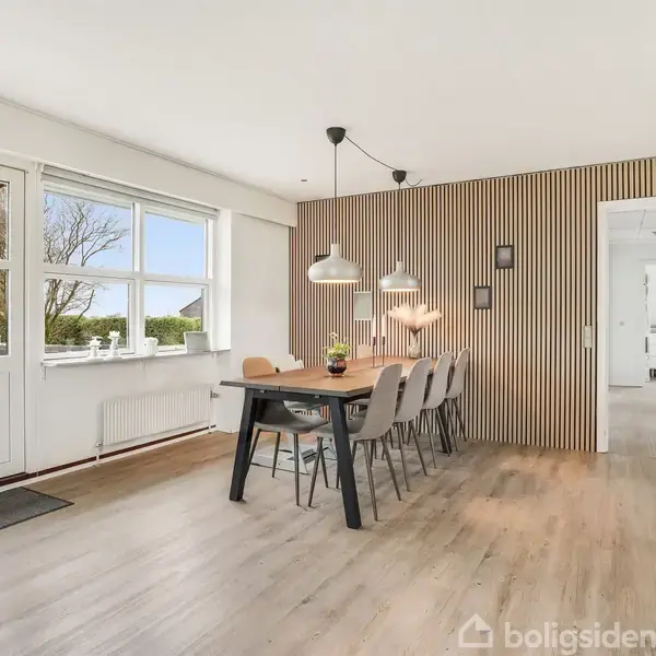 Et spisebord med flere stole står i et lyst rum med store vinduer og en dør til haven. To moderne lamper hænger over bordet, som står på et trægulv.