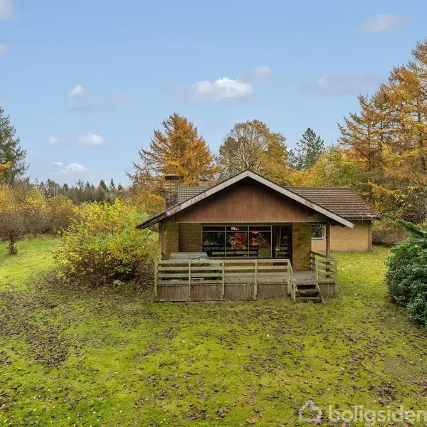 Et lille træhus med veranda står i en frodig skoveng, omkranset af efterårstræer. Græsplænen foran er dækket af faldne blade, og himlen er let overskyet.