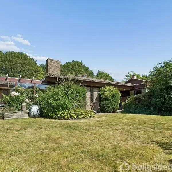Et moderne hus med fladt tag står omgivet af frodig have og buske, under en blå himmel. Græsplænen danner en stor del af forhaven, mens skoven kan ses i baggrunden.