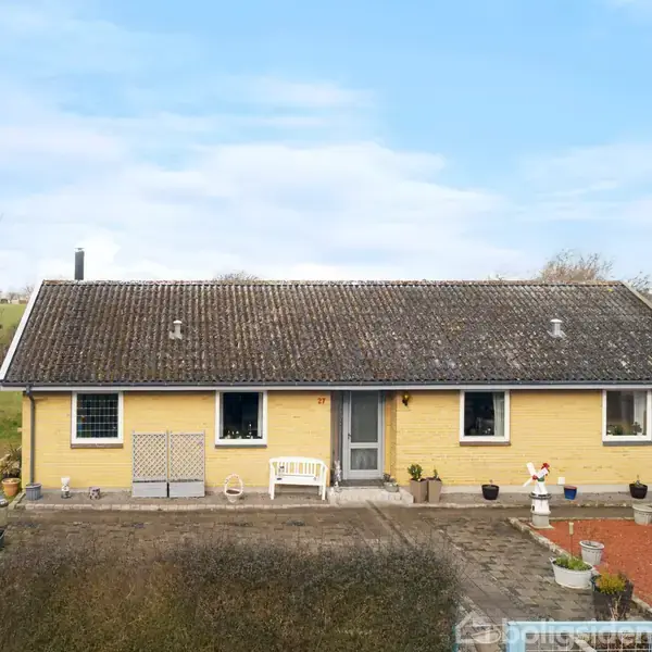 Et gult hus med et gråt tag står på en flisebelagt gårdsplads. En hvid bænk og potteplanter er foran huset. Baggrunden er en mark med træer.
