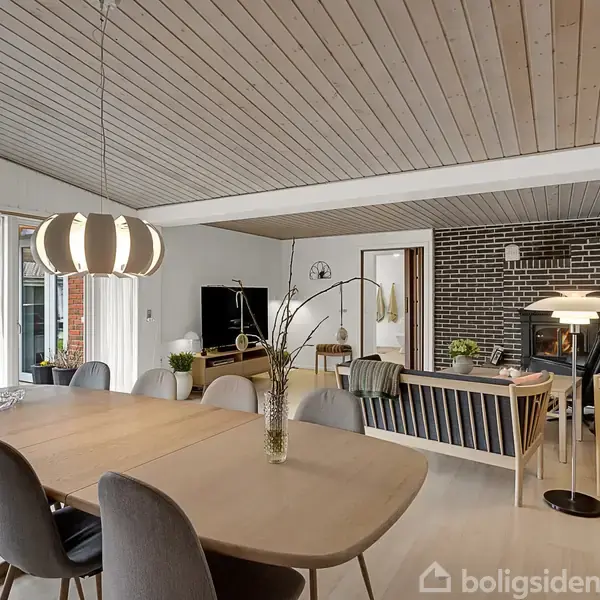 Et moderne, åbent spisestueområde med et langt træbord og grå stole. Et opholdsområde med pejs og sofaer ses i baggrunden. Lyse trævægge og loft skaber en hyggelig atmosfære.