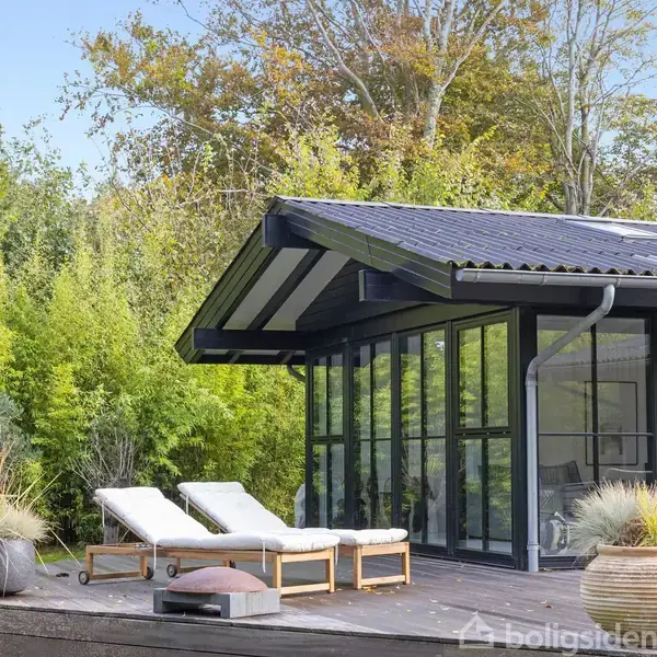 Et moderne sommerhus med store glasfacader står stille på en træterrasse. To liggestole står ved siden af krukker med planter, omgivet af frodig vegetation og træer i baggrunden.