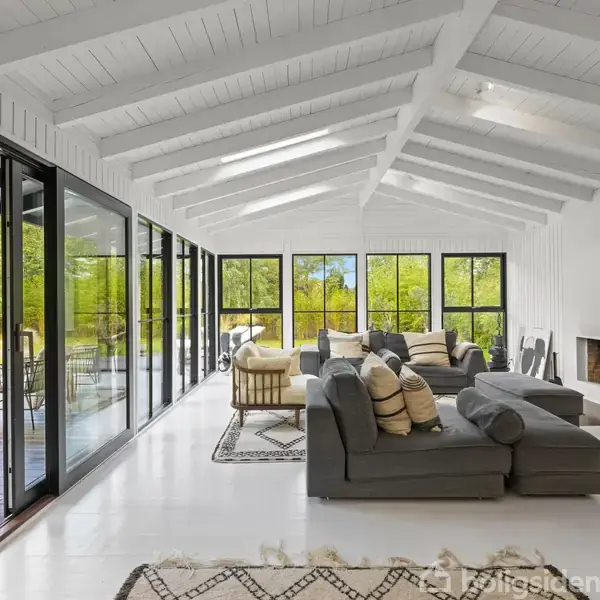 En grå sofa står i en lys stue med hvide trævægge og store vinduer med udsigt til en veranda og have. Et tæppe dekorerer det skinnende gulv. Pejs i hjørnet.