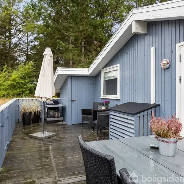 Terrassebord med stole står på en træterrasse, omkranset af et blåt sommerhus med hvid ramme. En parasol er slået op, omgivet af natur og træer i baggrunden.