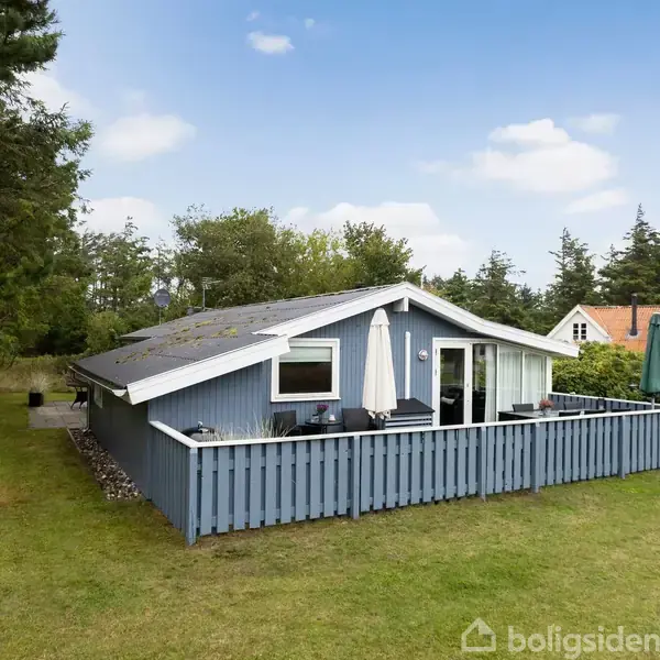 Et blåmalet sommerhus med terrasse står omgivet af grønne træer og græs. På terrassen ses havemøbler og parasoller. Baggrunden inkluderer et hus med rødt tag og blå himmel med få skyer.