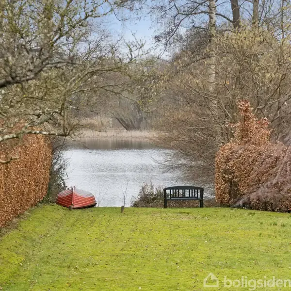 En rød robåd ligger på græsplænen ved siden af en sort bænk. I baggrunden ses en sø omgivet af træer og buske i vintertilstand.