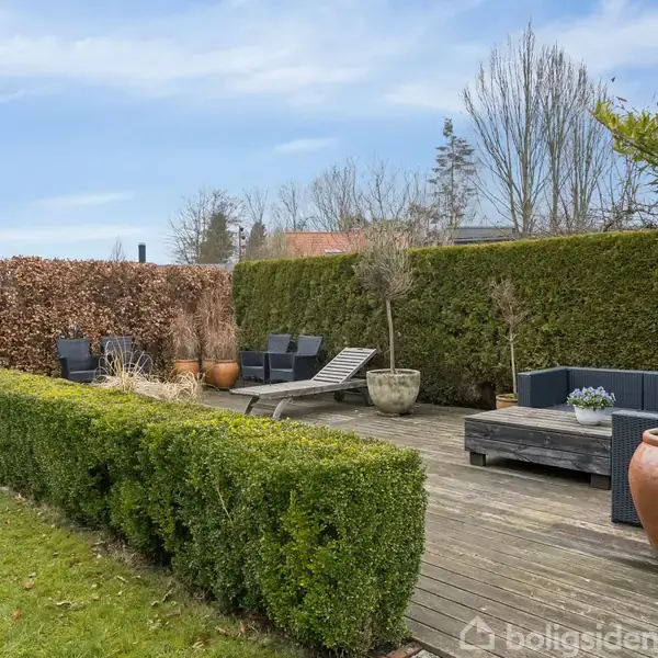 En terrasse med træmøbler og liggestole står omgivet af høje, tætte hække. Store potteplanter er placeret omkring området, og en græsplæne strækker sig foran terrassen.