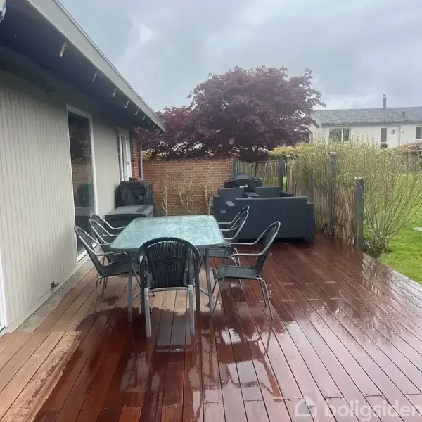 En udendørs terrasse med seks stole omkring et glasbord; møblerne er placeret på et trædæk. Omkring er der en murstensvæg, hække og et stort træ i baggrunden.