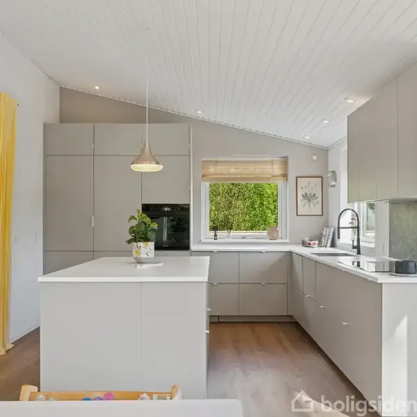 Køkken med grå skabe og hvid bordplade, hvor der står en plante. Skinnende vask ved et vindue med udsigt til grønne træer. Gule gardiner i baggrunden.