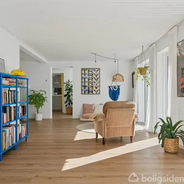 En stue med trægulv og blå bogreol fyldt med bøger til venstre. En beige stol står ved vinduerne, omgivet af planter og kunst på væggene.