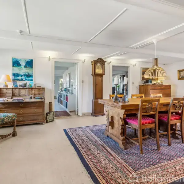 Træbord med seks stole står på et orientalsk tæppe i en rummelig stue. Omkring er der en kommode, et ur, og malerier på væggene. Store vinduer giver lys.