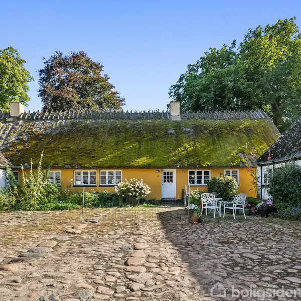 Gul bindingsværkshus med mosdækket stråtag står stille mellem to ejendomme. Stole og bord står til højre på gårdspladsen, omgivet af frodig vegetation og store træer i baggrunden.
