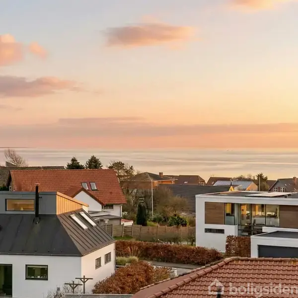Moderne huse med flade tage står stille i en rolig forstad, omgivet af lav bevoksning, mens solen går ned og farver himlen i bløde nuancer over en fjern vandhorisont.
