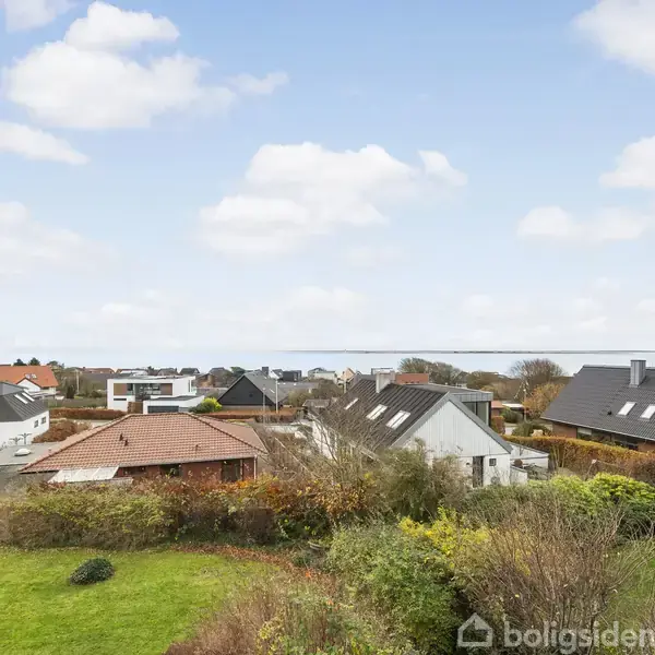 Flere huse med skrå tage står stille i et boligområde omgivet af velplejede haver og buske, mens en blå himmel med spredte skyer danner baggrunden.