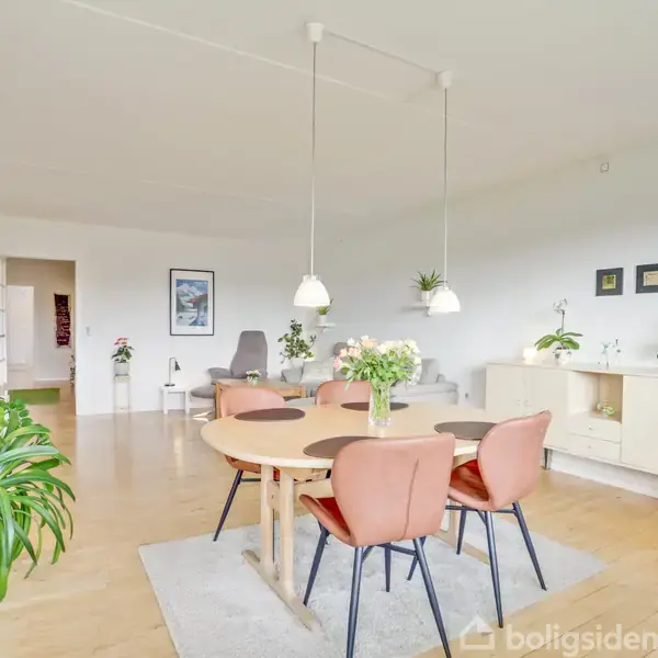 Et lyst spisestueområde med et rundt træbord omgivet af fire lyserøde stole. Bordet har en vase med blomster. Rummet har trægulv og vægge dekoreret med forskellige planter og billeder.