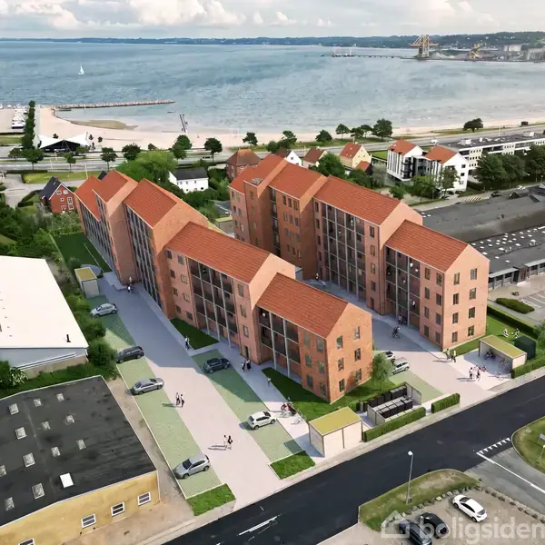 Et boligkompleks med røde tage og murstensfacader står stille nær en kystlinje. I baggrunden ses havet og en strand, omgivet af bygninger og veje i et byområde.