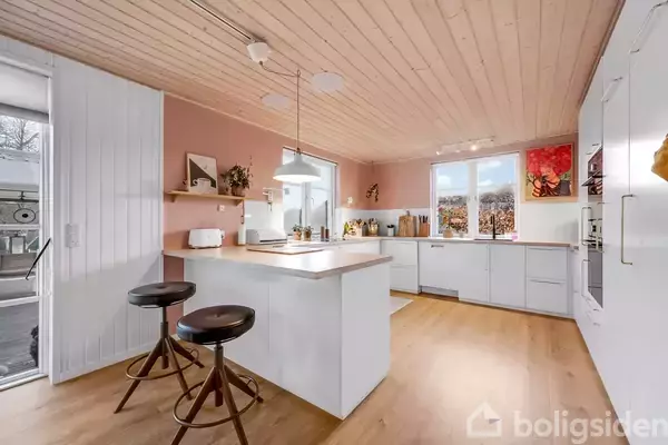 Et moderne køkken med en stor, hvid ø står i centrum. To barstole er placeret ved øen. Rummet har lyserøde vægge, træloft og vinduer med udsigt til naturen.