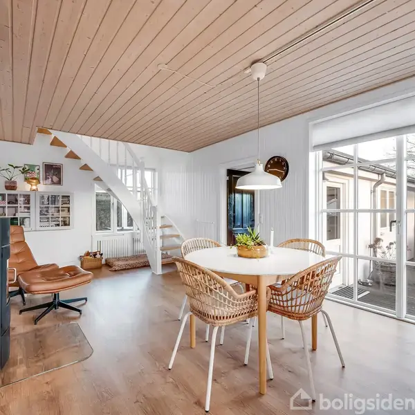 En moderne stue med spisebord, omgivet af firkantede stole, ved siden af store vinduer mod terrassen. En brændeovn står i hjørnet nær en trappe, der fører opad.