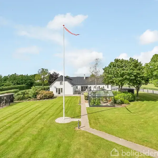 En hvid bygning med sort tag står i baggrunden. En flagstang med et rødt vimpel står i forgrunden. Omkringliggende græsplæne og træer skaber en rummelig have.