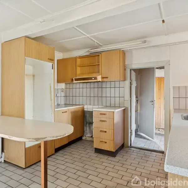 Et køkken med træskabe og fliser på væggene. Der er en hjørnevask, gasblus, og et lille spisebord. Værelset har lyse fliser på gulvet og en åben dør til højre.