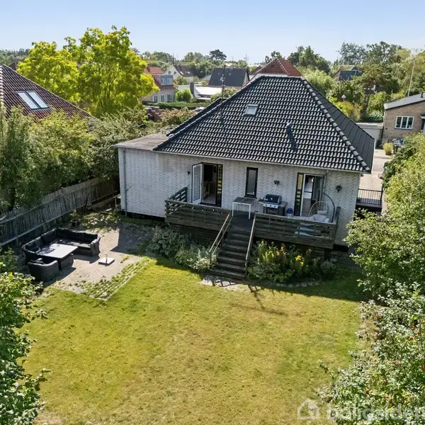 Et hvidt hus med sort tag står i en have. Huset har en terrasse med trappe og møbler. Haven er grøn og omgivet af træer og buske i et boligområde.